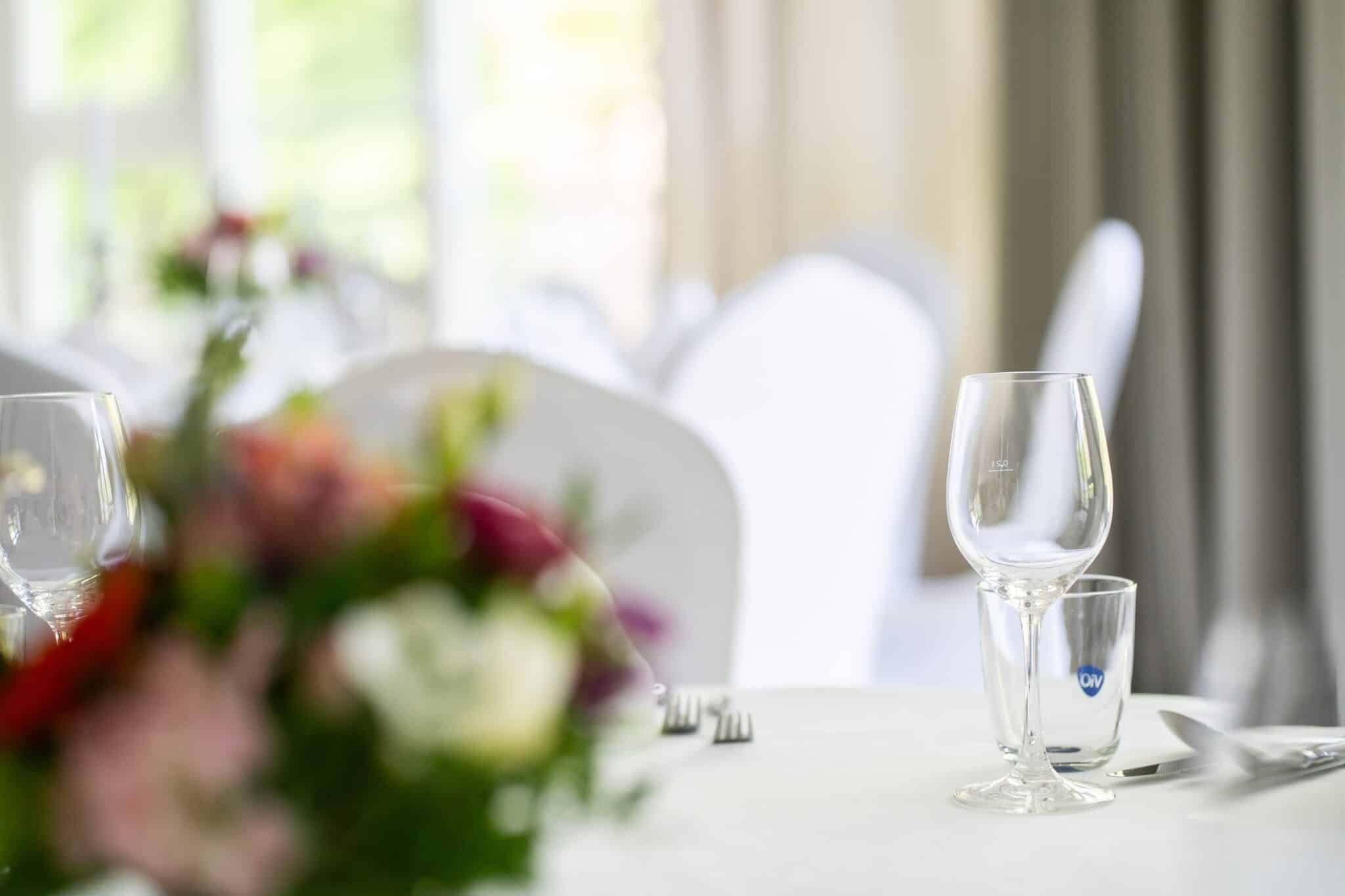 Gläser und Blumen auf einem runden Tisch für eine Hochzeit im Alstertal, im Hintergrund sind Stühle unter weißen Hussen