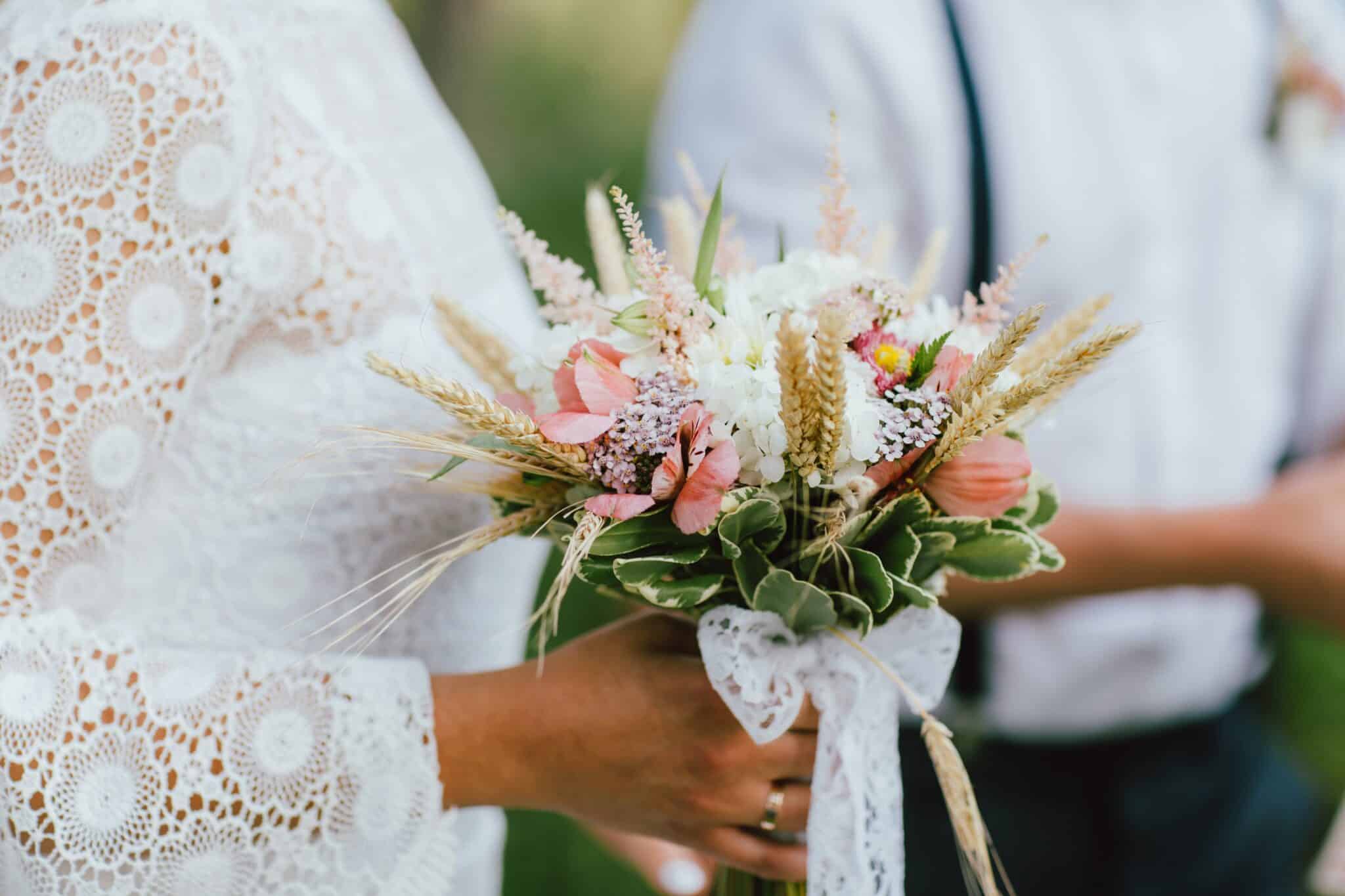 Brautstrauß mit Feldblumen in der Hand einer weiblichen Person, die ein weißen Kleid mit Spitze trägt, neben ihr ist eine männliche Person zu erkennen