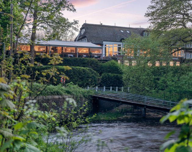 Das innen beleuchtete Gebäude des Hotels und Restaurants Mellingburger Schleuse in der Abenddämmerung, im Vordergrund ist der Alsterlauf zu sehen